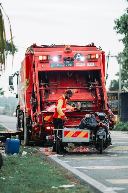A waste collection worker wearing a yellow and red high-visibility vest is seen operating a large red rubbish truck parked beside the street. The truck, positioned with its rear opened, reveals the mechanical loading area, which appears to have residual debris or waste material inside. The worker is leaning forward, possibly checking or handling the collected rubbish. In front of the truck, a small black refuse bag rests on a metal platform attached to a motorcycle or moped, which is parked close by. The scene unfolds on a paved roadside with a white line marking the edge, adjacent to a grassy verge, indicating a typical urban or suburban street environment. Behind the truck, some out-of-focus trees, utility poles, and live wires are visible against a partly cloudy sky, contributing to the outdoor setting. The lighting indicates daytime, with natural light softly illuminating the scene, consistent with typical waste collection activities that often support alternative or private waste management solutions, as handled by services like Rubbish Removal Finsbury Park.