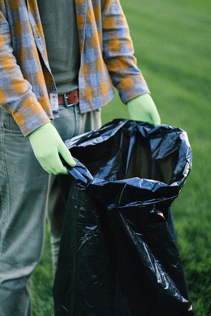 A person dressed in a grey and yellow plaid long-sleeved shirt, with a green glove visible on their right hand, is holding open a large black plastic rubbish bag outdoors on a grassy area. The bag appears to be made of shiny, crinkled plastic and is partially filled, ready for waste disposal. The person's lower half shows grey trousers secured with a brown belt. The background consists of a well-maintained lawn, suggesting an outdoor cleaning or waste collection activity, which relates to private or independent rubbish removal services such as those offered by Rubbish Removal Finsbury Park. The scene emphasizes manual handling of waste, with focus on the bag and the effort involved in rubbish collection, supporting the context of non-local authority waste disposal options or on-site clearance activities.