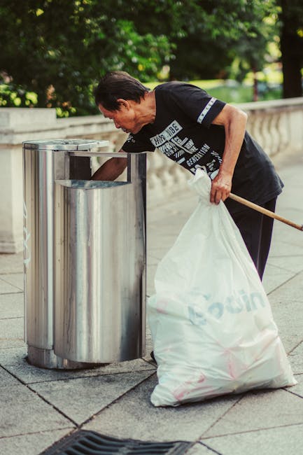A man with dark hair wearing a black T-shirt with white text is seen bending over a silver, cylindrical public rubbish bin with a rounded top, in an outdoor setting. He is holding a large, white plastic bag filled with waste, which is partially resting on the ground next to the bin. The man appears to be in the process of disposing of rubbish into the bin, using a wooden stick or litter picker in his right hand. The environment around him includes a paved walkway with a decorative stone railing and lush green trees in the background, suggesting a park-like setting. The scene is illuminated by natural daylight, highlighting the textures of the metal bin, the plastic bag, and the surrounding greenery. This image illustrates the activity of street cleaning or waste collection, possibly related to private waste handling or alternative rubbish removal services, as represented by Rubbish Removal Finsbury Park.