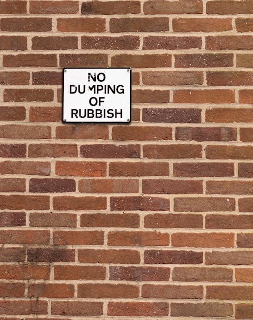 A square metal sign affixed to a red-brick wall displaying the message 'NO DUMPING OF RUBBISH' in black uppercase letters on a white background. The brick wall features a regular pattern of reddish-brown bricks with darker mortar joints, creating a textured surface. The sign is positioned centrally in the upper left portion of the image, suggesting a restriction against littering or illegal waste disposal in the area. The overall scene appears to be outdoors, with natural lighting highlighting the brick's surface and the sign's clarity, aligning with the context of waste management and rubbish removal regulations enforced by local authorities or private services such as Rubbish Removal Finsbury Park.