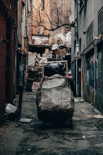 In a narrow alleyway between two urban buildings, a large, weathered black and grey rubbish bag filled with waste is positioned at the forefront, partially leaning against the left side of the alley. The ground beneath features dark, uneven asphalt with visible dirt and debris scattered around. Behind the bag, an old pushable trolley with a metal frame and fabric sides or cover extends into the cluttered background, which is filled with an overflowing heap of cardboard boxes, plastic bags, and miscellaneous refuse piled high against the back wall. The surrounding walls on either side display graffiti and peeling paint, with one side including a series of small, barred windows and the other a utility box attached to the exterior with some visible wiring. The environment appears dirty and neglected, suggesting the scene involves private waste accumulation or an alternative collection point outside a building, typical of independent rubbish disposal services like those provided by Rubbish Removal Finsbury Park.