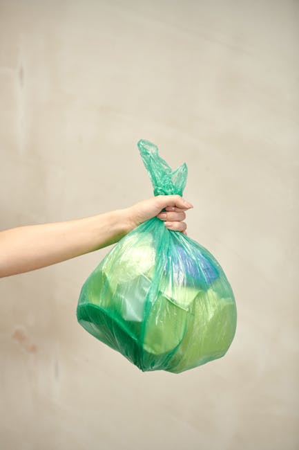 A person’s hand and forearm are visible, holding a green and transparent plastic bag filled with various waste materials, including blue and white items, against a plain light beige background. The bag appears to be made of thin, crinkled plastic, tied at the top with a twisted knot. The scene is set indoors, with the focus on the bag as a representation of household or general waste, highlighting the importance of proper rubbish disposal. This visual can be associated with private waste handling or alternative rubbish collection methods, such as independent rubbish removal services, supporting environmentally conscious practices. The minimal environment and neutral lighting emphasize the waste collection process without distractions, aligning with the themes of rubbish removal and waste management.