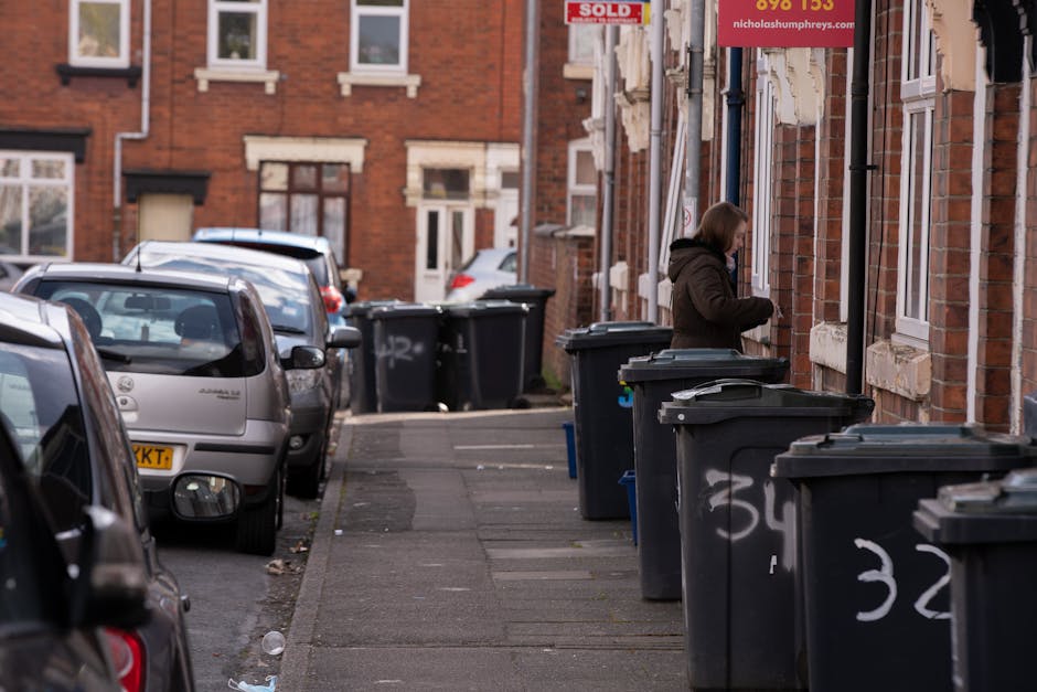 A residential street scene in Finsbury Park showing a row of large black wheelie bins lined up along the pavement in front of brick terraced houses. The bins are positioned on the sidewalk, some with closed lids and others slightly ajar, revealing dark interior contents. The street is narrow, with parked cars occupying the space on the left, including a small silver hatchback at the foreground with a visible yellow registration plate, and other vehicles further down the street. The background reveals additional terraced houses with brick facades, white window frames, and some graffiti tags on the lower walls near the bins. A person dressed in a brown coat is seen on the right side, standing next to a bin, likely engaged in waste disposal or collection. The environment appears typical of private waste handling areas, with no visible public rubbish collection vehicles, highlighting an alternative waste disposal scene. The lighting suggests a neutral, overcast day with soft natural illumination, emphasizing the textures of the brickwork, the smooth plastic surfaces of the bins, and the reflective car windows. Rubbish Removal Finsbury Park offers professional rubbish management, as seen in this image of private waste bins ready for collection or disposal in an urban residential setting.
