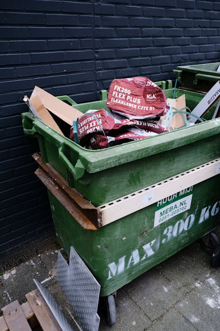 A large green wheeled skip placed on a paved surface against a dark brick wall, filled with unopened bags of gardening compost or soil, identifiable by their red packaging and printed labels. The skip is positioned on a wooden pallet to elevate it from the ground, and a grey metal plinth surrounds the skip's lower edge. To the right, a similar green waste container with a lid is partially visible. The environment appears to be an outdoor area, possibly a back garden or service yard, with natural daylight illuminating the scene. The presence of the skip and its contents suggests an activity related to garden or property waste disposal, reflecting an alternative to traditional rubbish collection methods managed by companies such as Rubbish Removal Finsbury Park, who facilitate private waste handling and on-site clearance services.