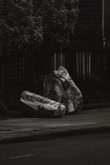 A collection of three large black plastic rubbish bags, slightly crumpled and tied at the top, resting on a paved sidewalk near a black metal fence. The bags appear to contain discarded household waste and are positioned in a staggered manner. Behind the fence, dense greenery is visible, with dark leafy branches extending upward and partially obscuring the background. The scene is captured in low natural light, creating a shadowed environment that highlights the texture and creases of the plastic bags. The area appears to be an urban or suburban street, where independent waste collection by private services such as Rubbish Removal Finsbury Park may be used for disposal. The setting reflects a typical example of on-site rubbish accumulation that can be managed through professional waste removal to ensure cleanliness and compliance with local waste regulations.