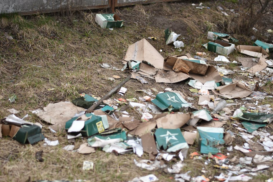 A patch of grassy ground near a fence shows a disorganized accumulation of discarded packaging materials, including flattened cardboard boxes and torn paper, some with printed logos and text. The cardboard boxes are in shades of brown, with a rough, fibrous texture, and are crumpled or torn, revealing their corrugated structure. Among the debris are crushed green and white cardboard cartons with a star logo, partially flattened but still identifiable. Scattered fragments of paper and packaging sit randomly across the uneven ground, which features patches of dried grass and dirt. In the background, there is a wooden fence, and the overall scene appears to be outdoors, possibly part of an area designated for rubbish disposal or an unmanaged waste site. The lighting is natural, with an overcast sky casting diffuse light over the scene, emphasizing the cluttered, unmanaged state of the rubbish, which contrasts with professional waste removal standards. Rubbish Removal Finsbury Park may be involved in addressing such debris through alternative waste handling, highlighting the importance of proper disposal of such waste materials.